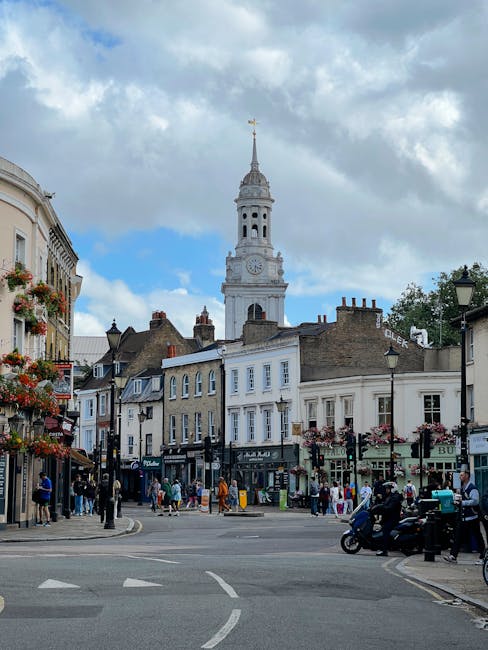 A street scene in Roman Road Market, Bow, showing a row of shops and pedestrians on the pavement. In the background, there is a historic white clock tower with a spire, set against a partly cloudy sky. The foreground features a road with vehicles, including a motorcycle and a scooter, waiting at a junction. On the left side, there are building facades with flower baskets and shopfronts, while the right side displays additional shops and street furniture. This image captures the busy atmosphere of a typical street in Bow, with ongoing home relocation and furniture transport activities supported by Bow Removals, illustrating the logistics involved in house removals and moving services in the area.