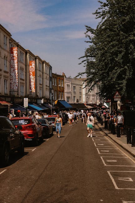 A busy street scene on Roman Road Market with pedestrians walking along the pavement and browsing shopfronts. Parked cars line both sides of the road, with some vehicles partially visible in the foreground. On the left, there are multi-storey buildings featuring retail shops and market stalls, some of which are covered with awnings and banners. People are seen carrying shopping bags and engaging with market vendors. In the background, a large tree provides shade, and the sky is partly cloudy with patches of blue. The setting reflects an active urban environment suitable for house removals and home relocation services, with potential opportunities for moving companies like Bow Removals to coordinate furniture transport and packing activities in lively market areas.