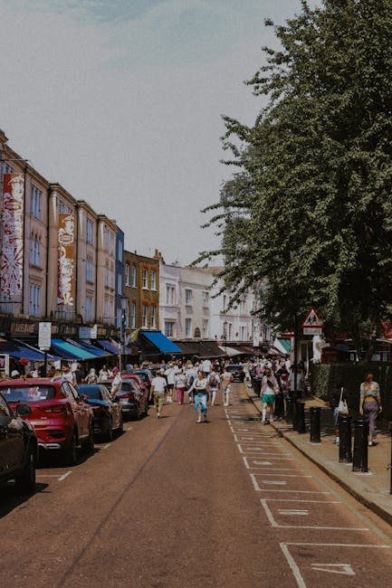 A street scene in Roman Road Market, Bow, showing a row of shops and pedestrians on the pavement. In the background, there is a historic white clock tower with a spire, set against a partly cloudy sky. The foreground features a road with vehicles, including a motorcycle and a scooter, waiting at a junction. On the left side, there are building facades with flower baskets and shopfronts, while the right side displays additional shops and street furniture. This image captures the busy atmosphere of a typical street in Bow, with ongoing home relocation and furniture transport activities supported by Bow Removals, illustrating the logistics involved in house removals and moving services in the area.